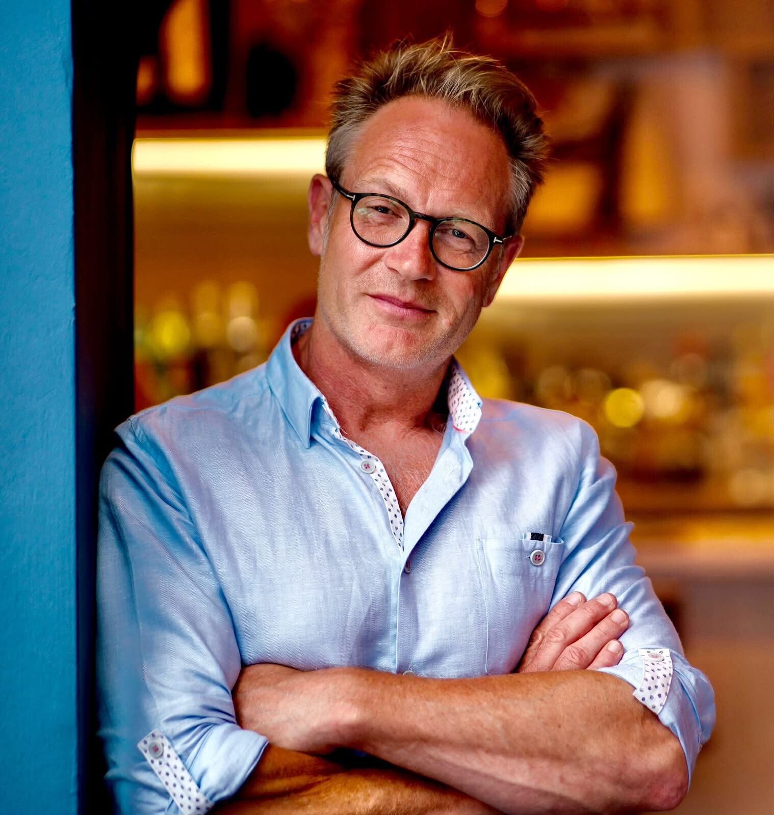Will Phillips, arms folded, in a pale blue linen shirt in front of the back bar at The Priory